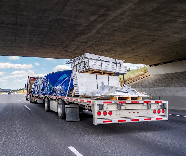 Semi truck transporting covered and tightened cargo on flat bed trailer on road under bridge.