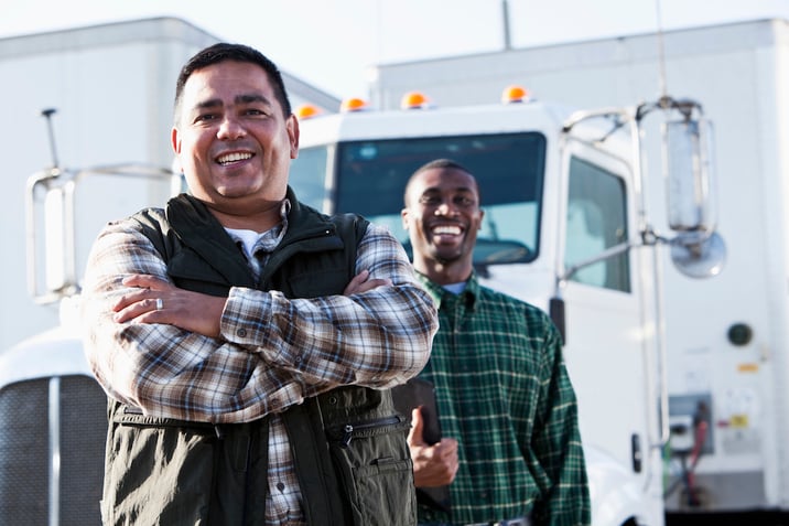 Two male truck drivers smiling in front of white semi truck in background.