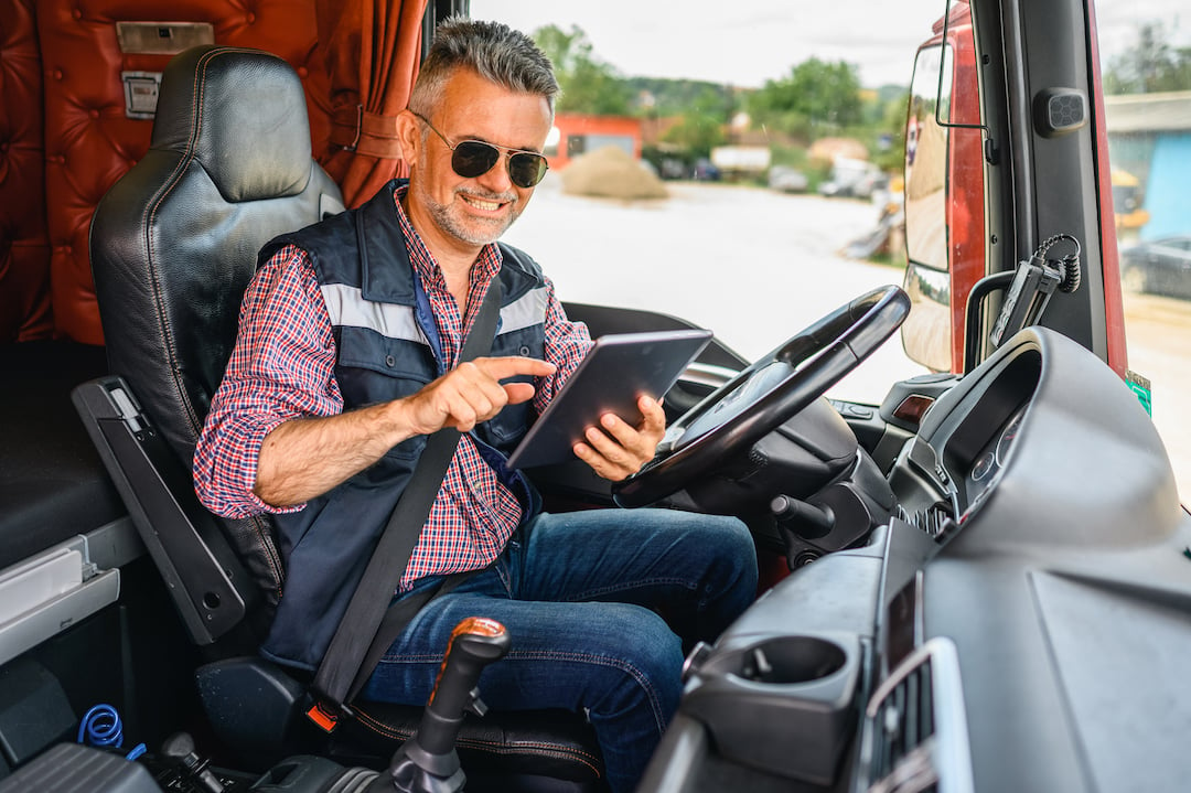 Old male truck driver sitting in truck cab using tablet.
