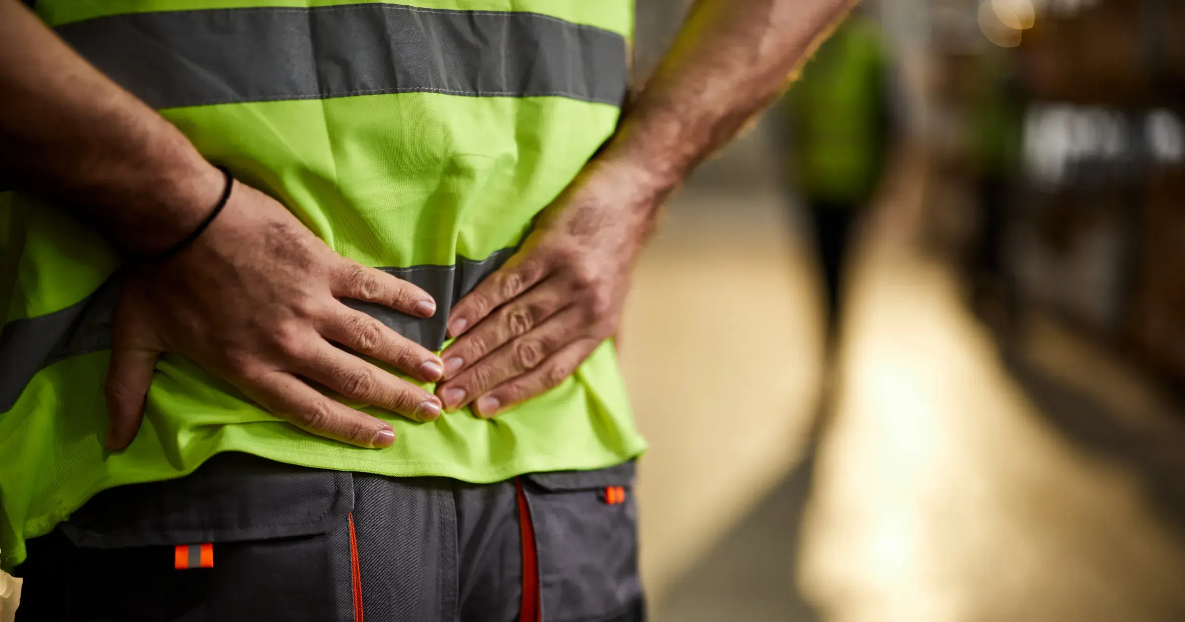 Warehouse worker in a high-visibility vest holding their lower back, indicating pain or injury, with a blurred background of an industrial setting.