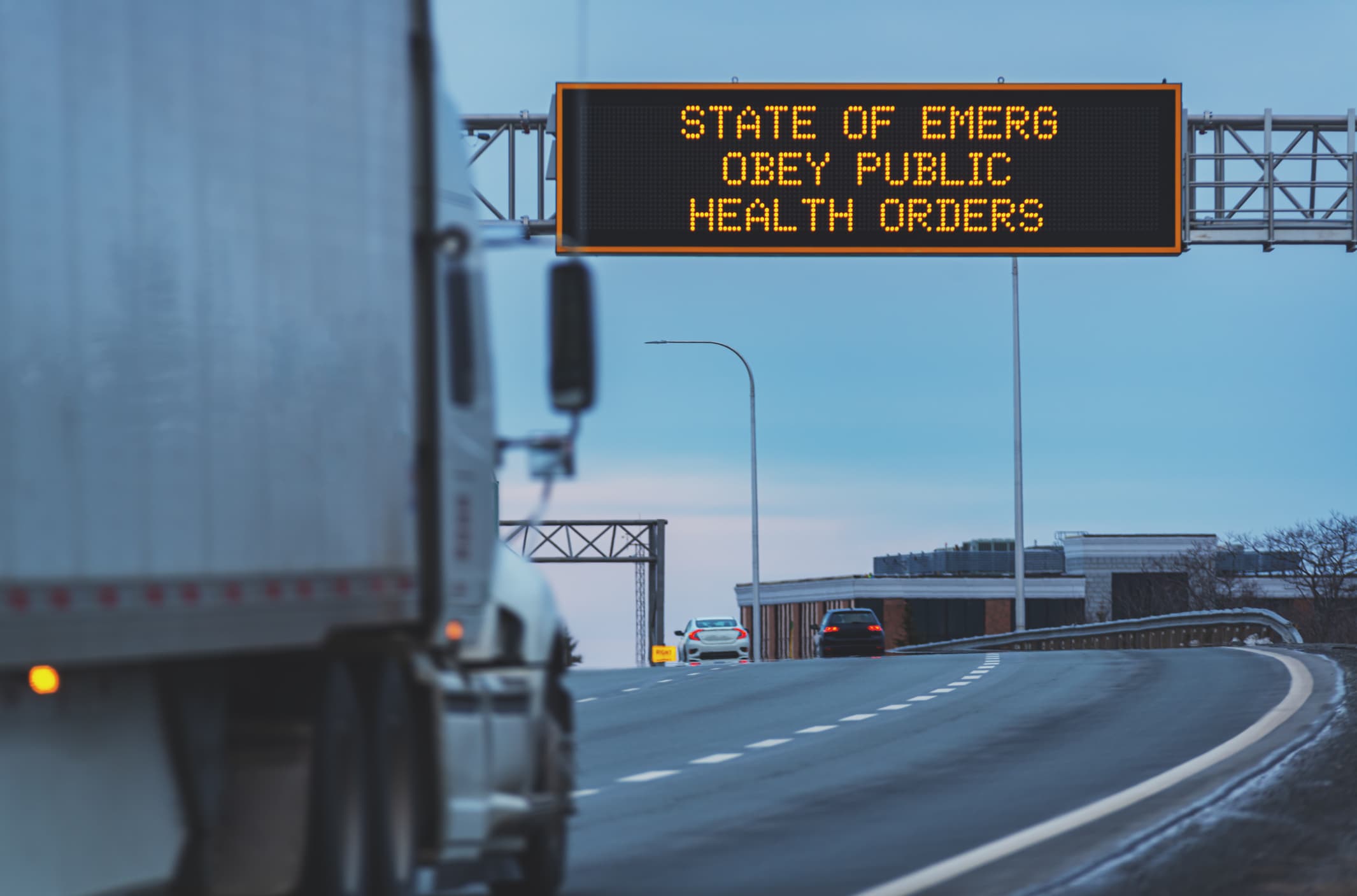 a tractor trailer driving down a highway under a sign that reads state of emergency obey public health orders
