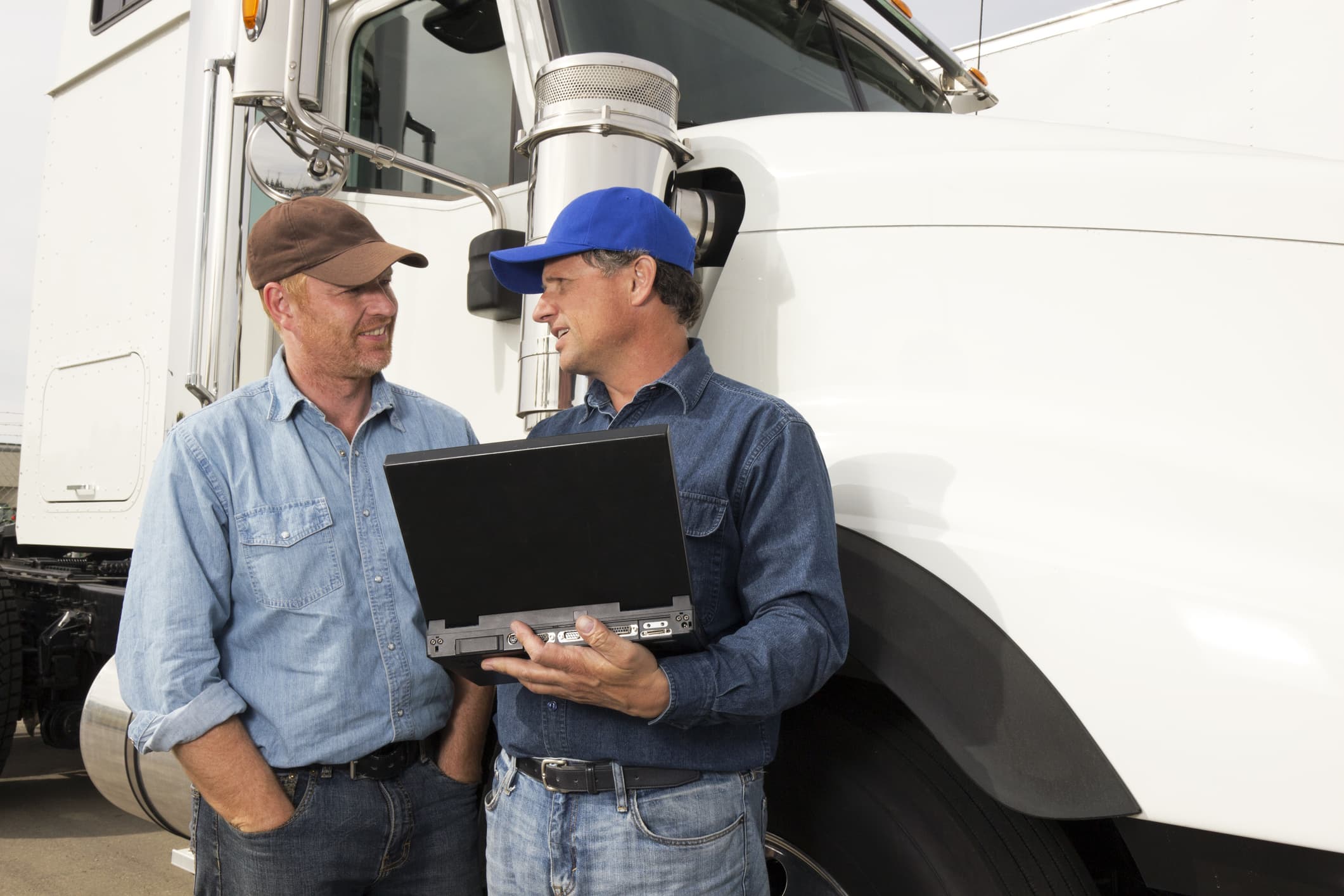two truck drivers standing next to a truck talking and holding a computer 